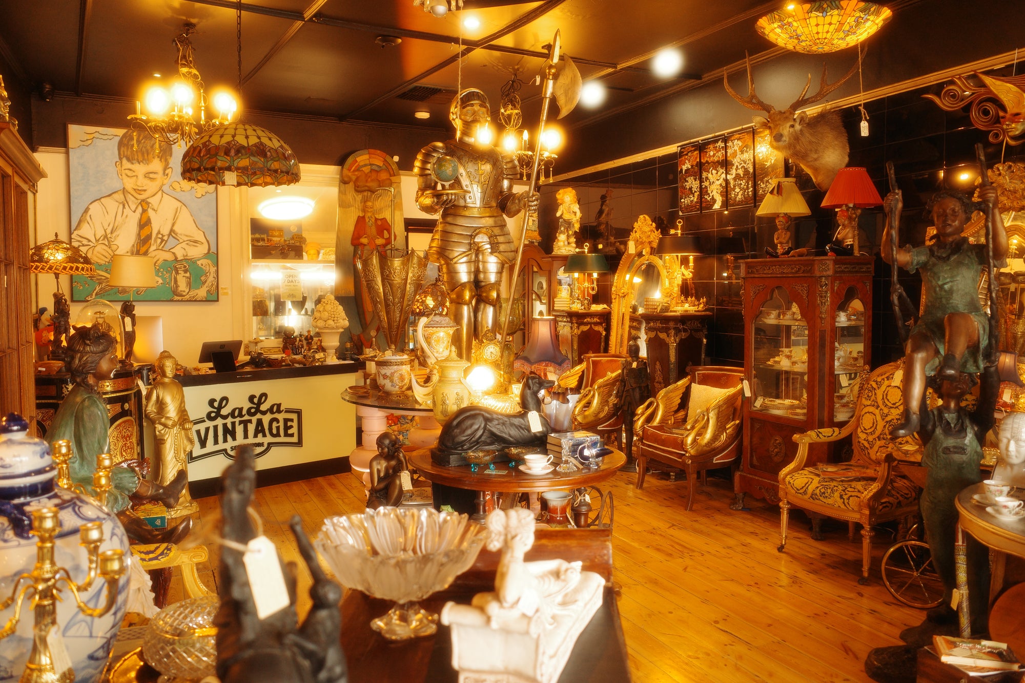 A beautiful eclectic antique shop filled with decorative items, including a large fibreglass knight statue, chandeliers, victorian furniture, and a sign reading 'La La Vintage'.