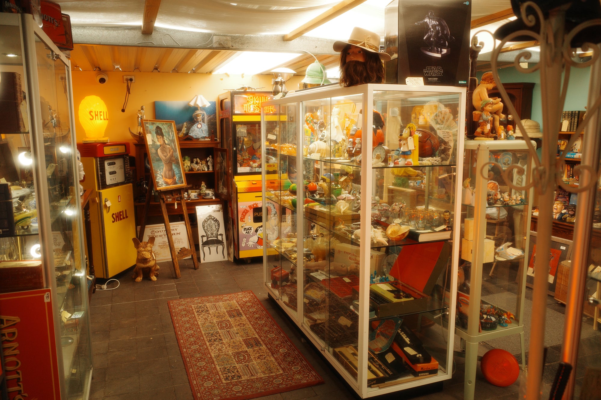 A cluttered vintage shop interior with multiple glass display cases filled with toys, figurines, and memorabilia. A vintage Shell petrol pump refrigerator stands on the left, an original Plucka Duck claw machine with neon next to it, and a patterned Persian rug is on the floor in the center.
