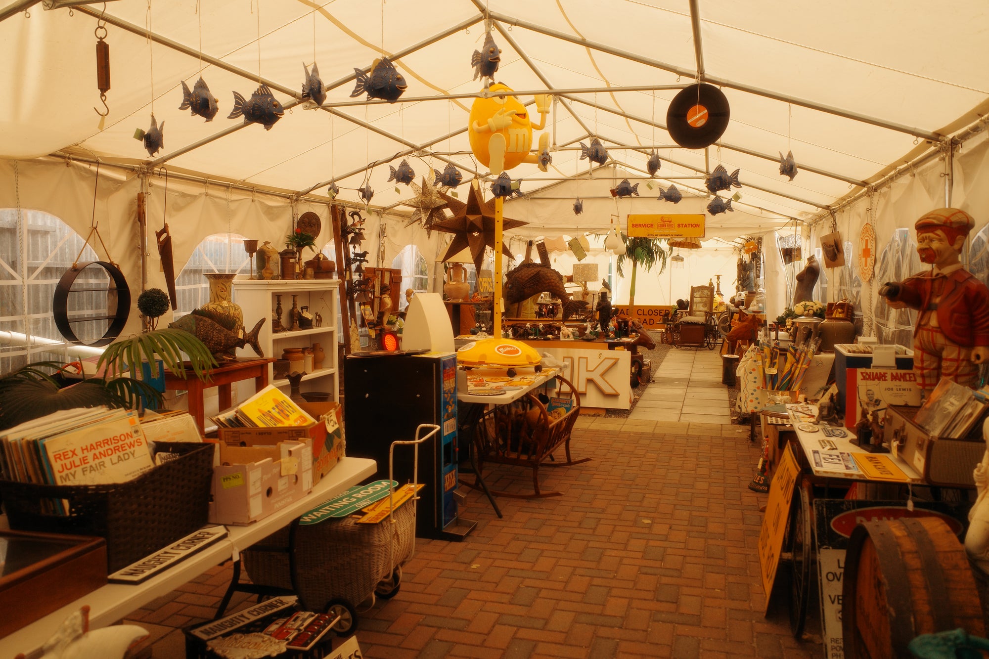 A long room under a large white event tent filled with assorted vintage and eclectic merchandise, including fish-shaped ornaments, record players, shelves, baskets, and display tables on a brick floor.