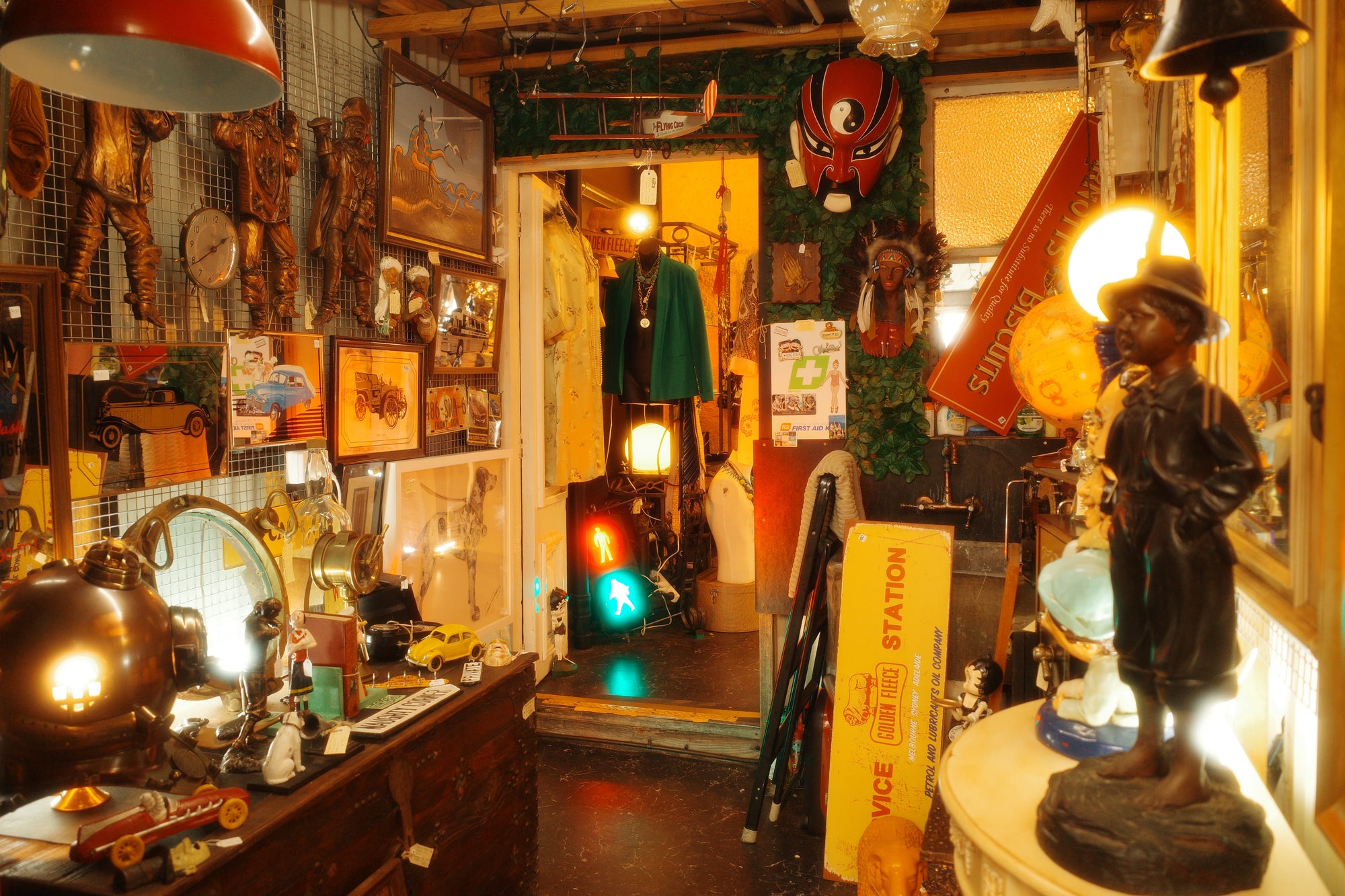 A cluttered vintage antique shop interior with warm lighting, displaying a collection of items including a brass helmet, unique figurines, framed pictures and bar mirrors, and cast iron models and signs.