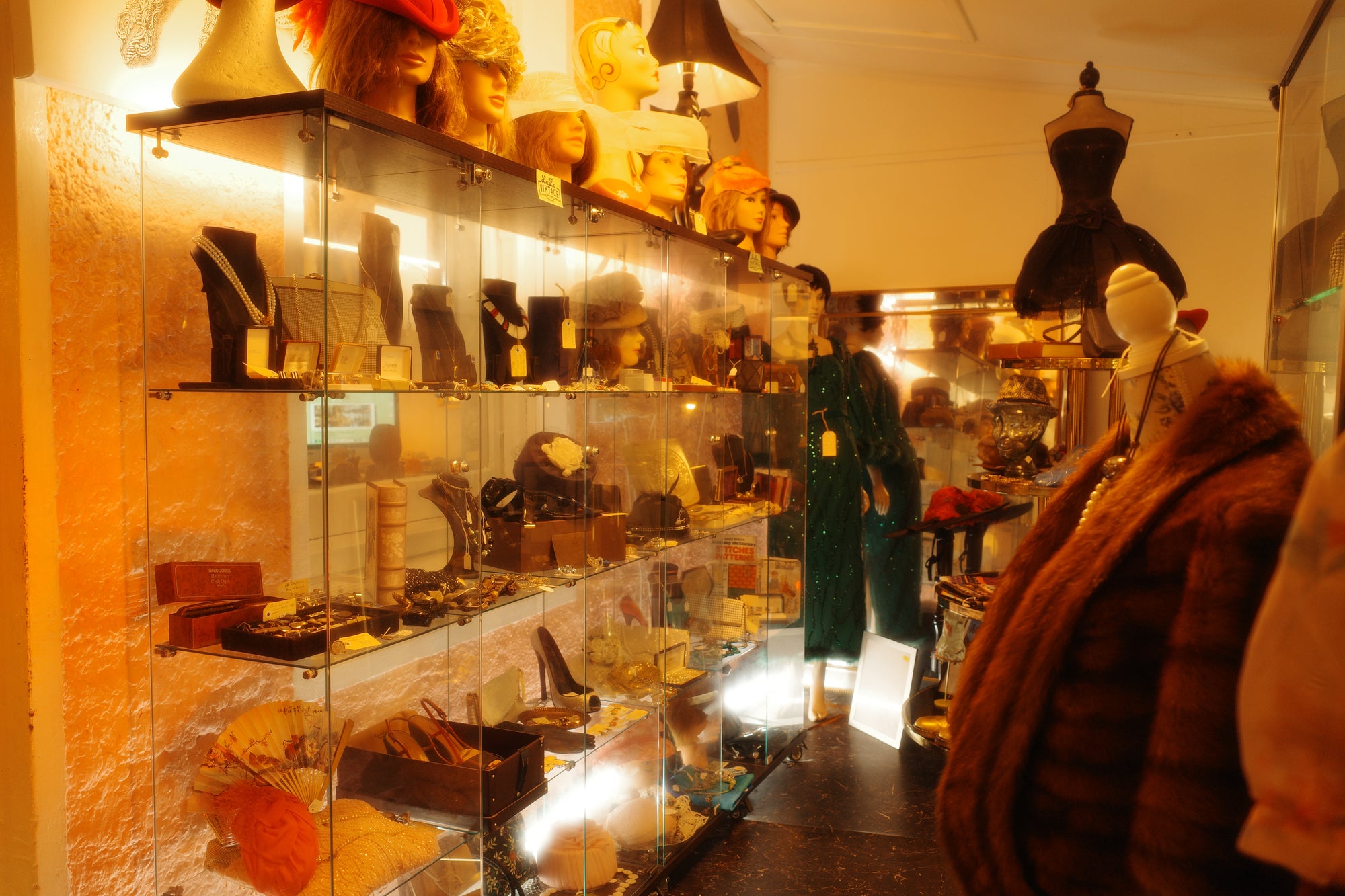 A warm, cozy vintage-style boutique interior with glass display cases filled with exotic fashion accessories and jewellery, mannequins wearing garments, and a fur coat in the foreground.