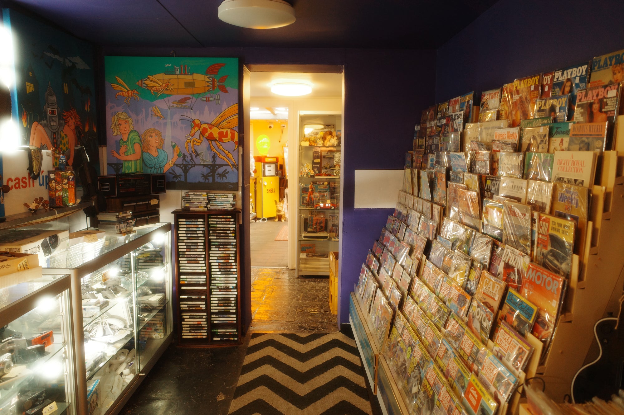 An organised antique shop retail interior with purple walls, a glass display cabinet of countless retro video games and vintage cameras on the left, a huge display of hundreds of vintage magazines and comics on the right, and a doorway in the back leading into many more rooms.